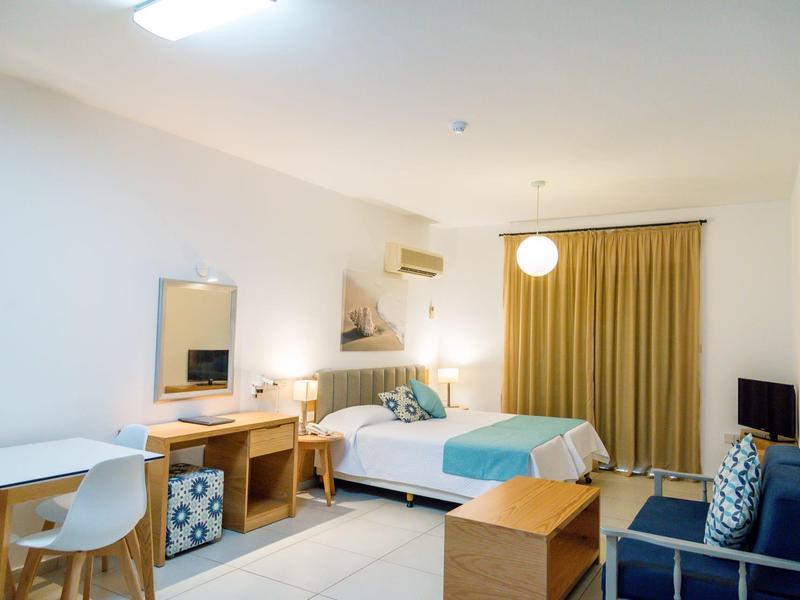 Helles Hotelzimmer mit Doppelbett, Schreibtisch, Sessel, TV und großen Vorhängen, schlicht eingerichtet.