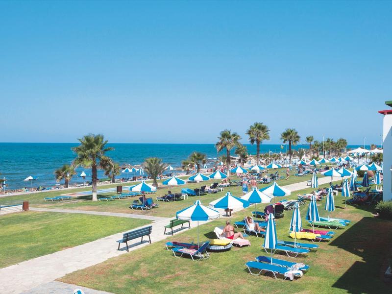 Strandpromenade mit Liegestühlen, blau-weißen Sonnenschirmen und Palmen am Meer unter blauem Himmel.