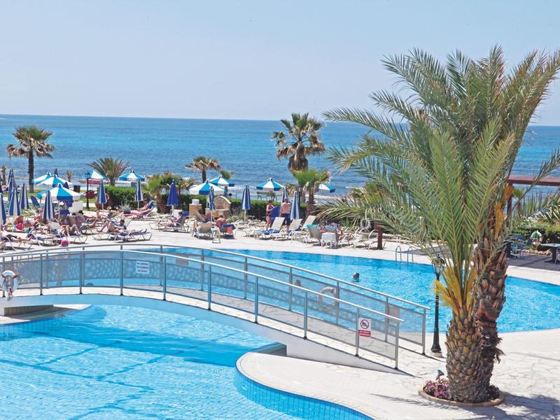 Großer Pool mit Brücke, Palmen und vielen Liegestühlen am Meer bei klarem Himmel.