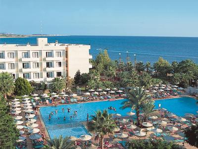 Großer blauer Hotelpool mit Sonnenschirmen, Palmen und weißem Gebäude am Meer im Hintergrund.