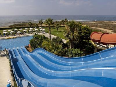 Mehrere blaue Wasserrutschen führen in einem Wasserpark mit Pool und Blick aufs Meer.