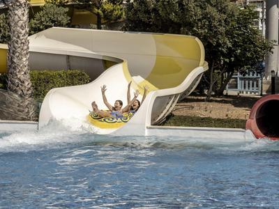 Drei Personen rutschen lachend eine gelbe Wasserrutsche hinunter in einen Pool.