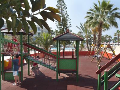 Kinder spielen auf einem Spielplatz mit Rutschen, Schaukeln und Klettergerüsten unter Palmen.