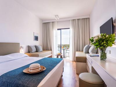 Chambre d'hôtel lumineuse avec lit double, canapé, balcon et table avec des fleurs.
