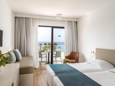 Modernes Hotelzimmer mit Doppelbett, Balkon, Meerblick, weißer Wand und hellen Holzmöbeln.