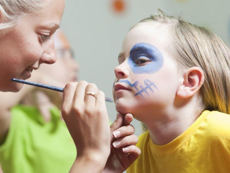 Eine Frau malt einem Kind mit bunter Gesichtsbemalung die Nase in einem hellen Raum.