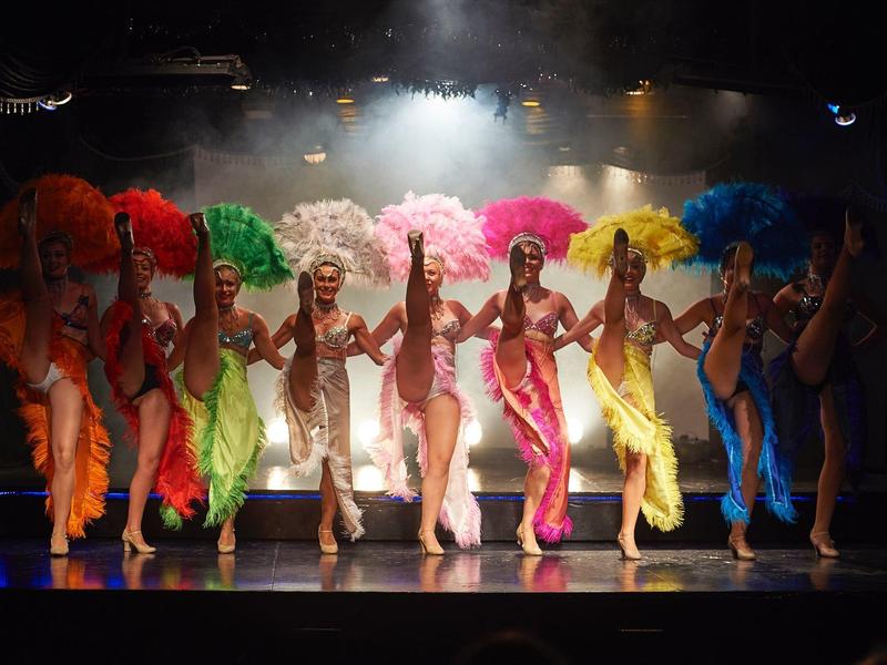 Sieben Tänzerinnen in bunten, federreichen Kostümen stehen auf einer Bühne unter hellem Licht.