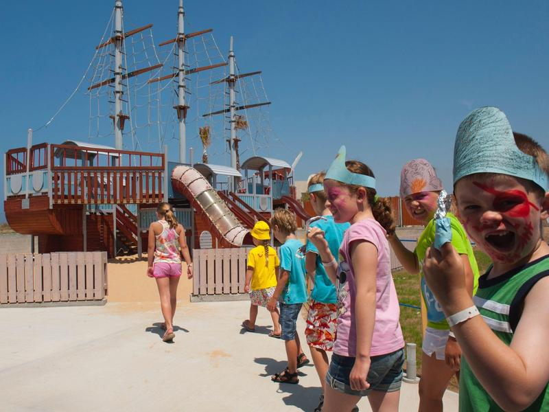 Kinder spielen im Freien vor einem großen, hölzernen Schiff mit Mast und blauem Himmel.