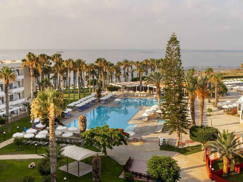 Großer Hotelpool mit Palmen, Liegen und weißen Gebäuden, im Hintergrund das Meer bei klarem Himmel.