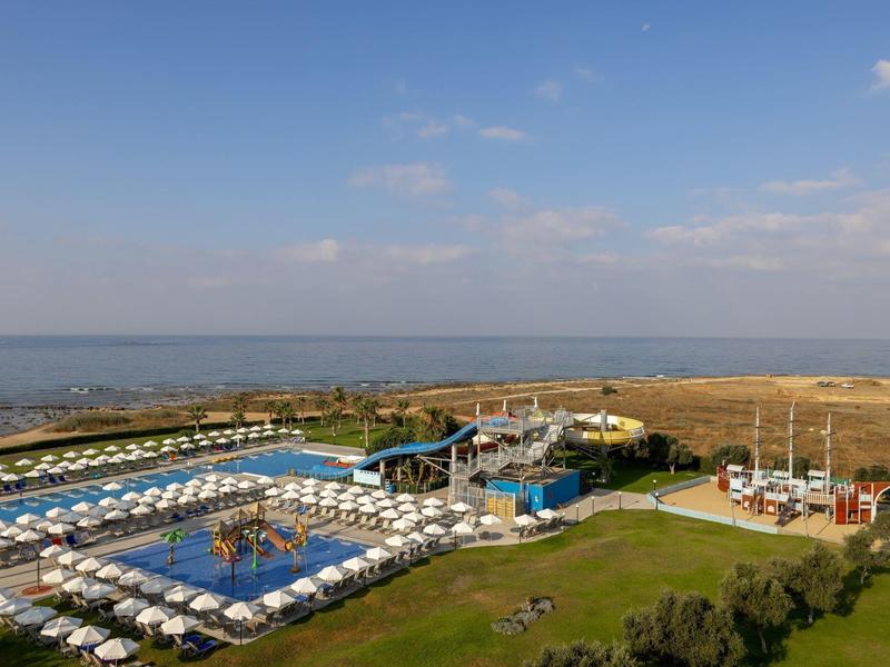 Großer Pool mit weißen Liegestühlen, grünem Rasen und Blick auf ruhige Meeresküste unter blauem Himmel.