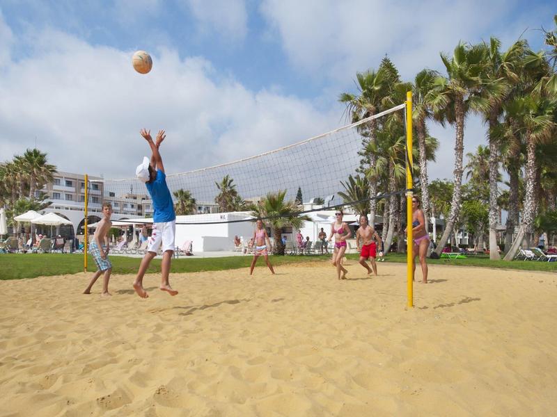 Mehrere Personen spielen Volleyball auf einem Sandplatz mit Palmen und Gebäuden im Hintergrund.