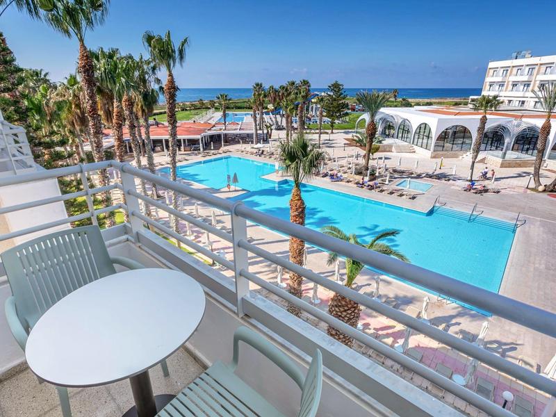 Blick von Balkon auf großen Pool mit Sonnenschirmen, Liegen, Palmen und blauem Himmel am Meer.