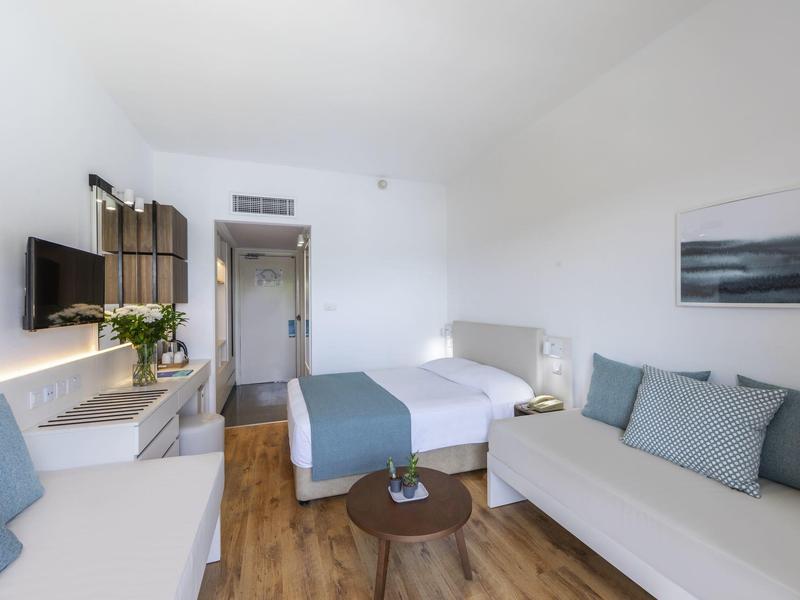 Moderner Hotelzimmer mit Holzboden, Doppelbett, Sofas, Wandbild und TV an weißer Wand.