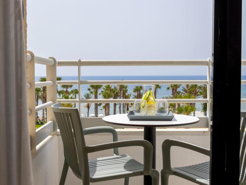 Balkon mit zwei Stühlen, Tisch, Getränk und Meer im Hintergrund bei sonnigem Wetter.