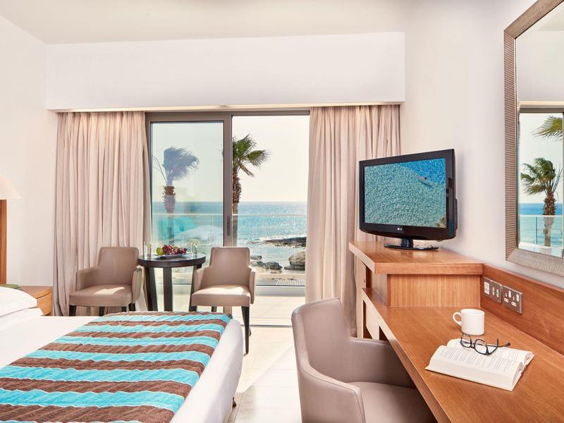 Helles Hotelzimmer mit Meerblick, Bett, Schreibtisch und Fernseher, Balkon mit Stühlen.