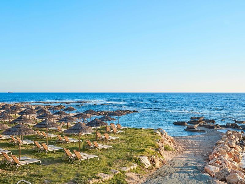 Strand mit Sonnenschirmen und Liegen neben einem Felsenweg zum Meer bei klarem Himmel