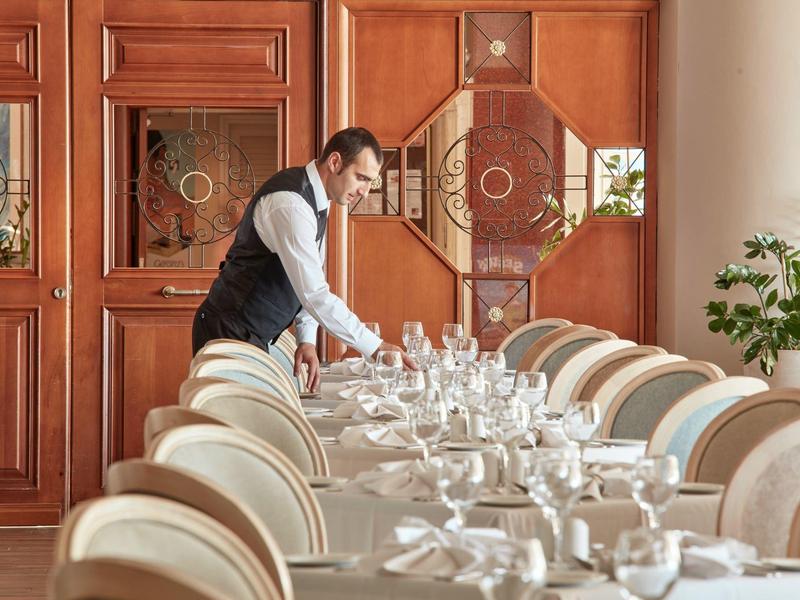 Ein Kellner richtet einen lang gedeckten Tisch in einem eleganten Speisesaal mit Holzverkleidung ein.