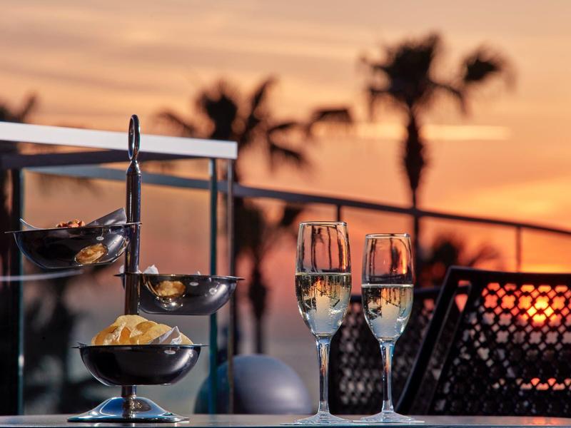 Champagnergläser und Snacks auf einem Tisch mit Palmen und Sonnenuntergang im Hintergrund.