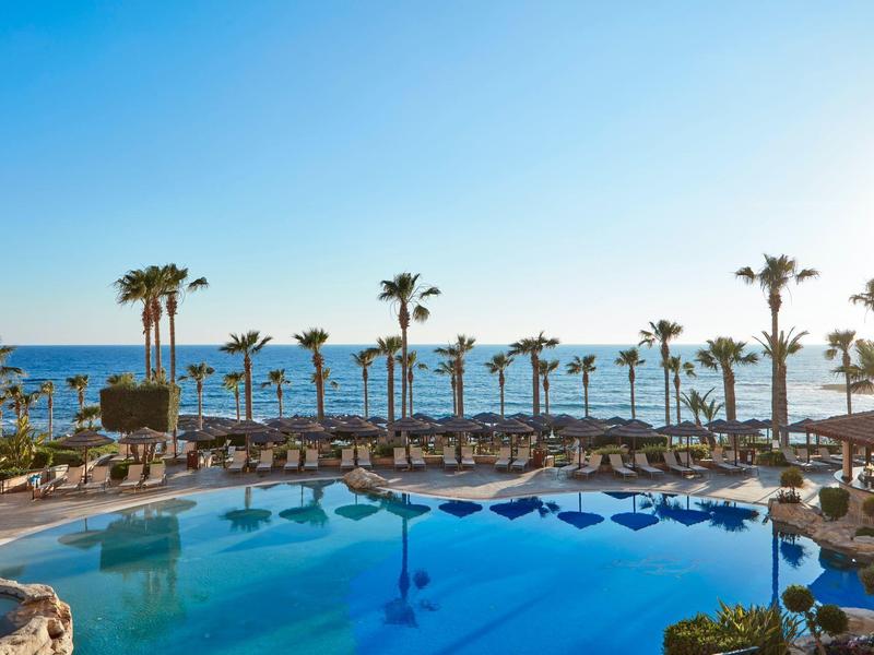 Großer Pool mit Sonnenschirmen und Palmen vor Blick auf das Meer bei klarem Himmel.
