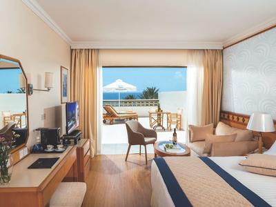 Helles Hotelzimmer mit Doppelbett, Terrasse, Blick auf Meer, Holzmöbeln und Sandfarbenen Vorhängen.