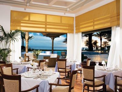 Helles Restaurant mit weißen Tischdecken, Holzmöbeln und Blick auf Palmen und Meer bei Sonnenuntergang.