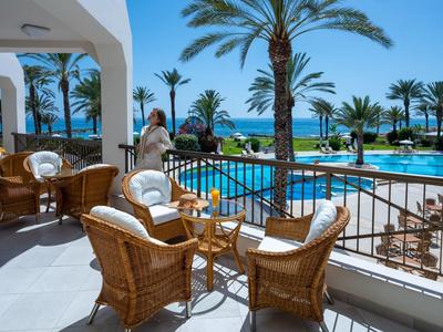 Terrazza con sedie di vimini con vista su palme, piscina e mare sotto un cielo limpido.