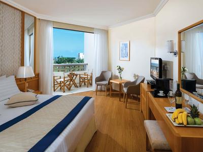 Camera d'hotel luminosa con balcone, pavimento in legno, zona soggiorno e TV a schermo piatto.