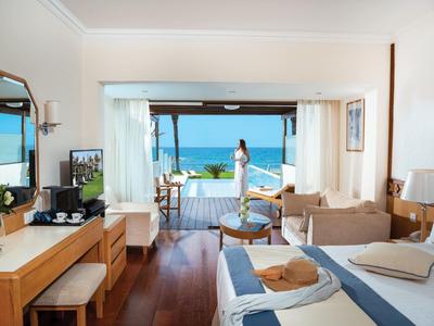 Camera d'hotel luminosa con vista mare e balcone, letto matrimoniale, divano e scrivania.