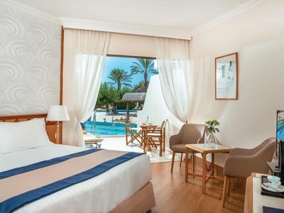 Camera d'hotel luminosa con letto grande, zona salotto e vista su piscina e palme.