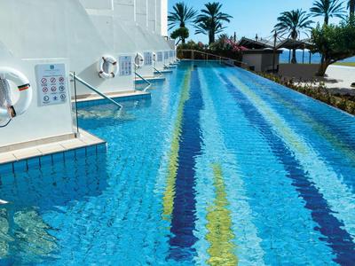 Piscina esterna lunga e stretta con strisce colorate e muri bianchi, circondata da palme.