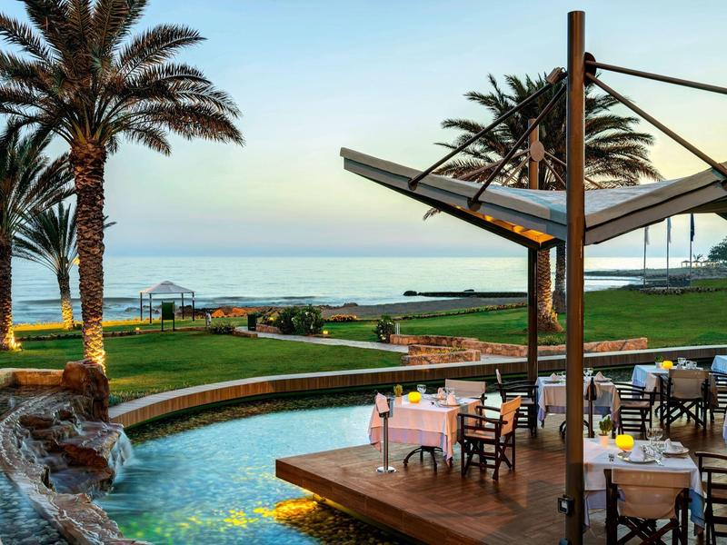 Strandrestaurant mit blauer Wasserfläche, grüner Wiese, Palmen und Meer im Hintergrund bei Tageslicht.