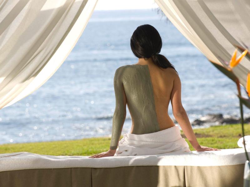 Frau mit Rückenmaske sitzt entspannt auf Massageliege, Blick aufs Meer, helle Vorhänge umrahmen Szene.