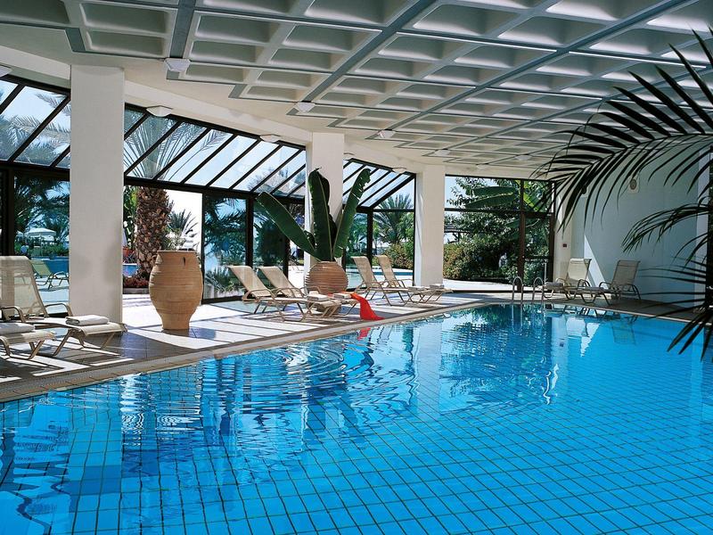 Hallenbad mit blauen Kacheln, Liegestühlen, großen Fenstern und üppigem Pflanzen-Dekor.