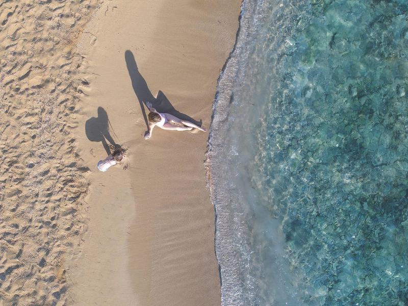 Zwei Personen liegen am sandigen Strand neben klarem, blauem Wasser mit sanften Wellen.