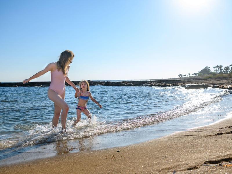 Zwei Mädchen spielen am sonnigen Strand im flachen Wasser, blauer Himmel und Sand sichtbar.