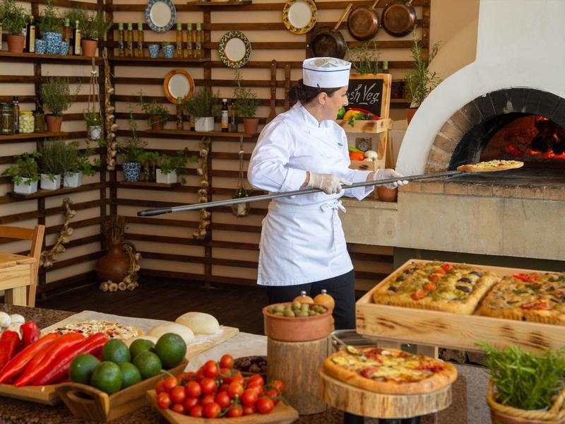 Ein Koch in weißer Uniform backt Pizza in einem traditionellen Holzofen, umgeben von frischem Gemüse.