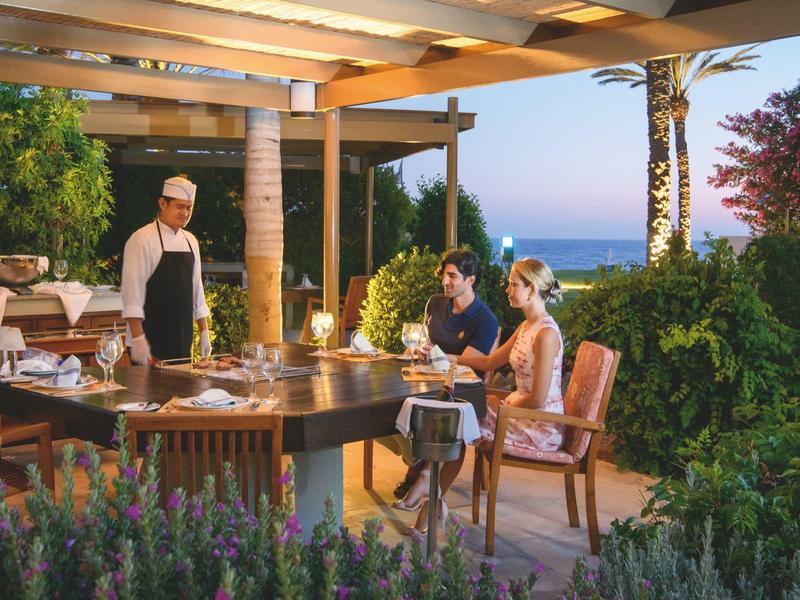 Abendessen auf einer Terrasse mit Meerblick, Kellner serviert Paar, umgeben von Pflanzen und Holzdeck.