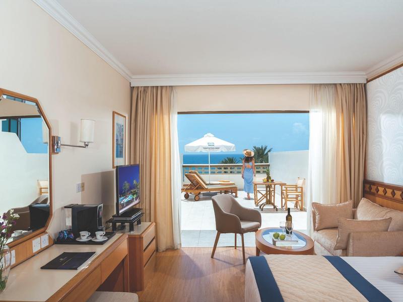 Helles Hotelzimmer mit Holzmöbeln, Sofa, TV und Blick auf Terrasse mit Meer und Sonnenschirm.