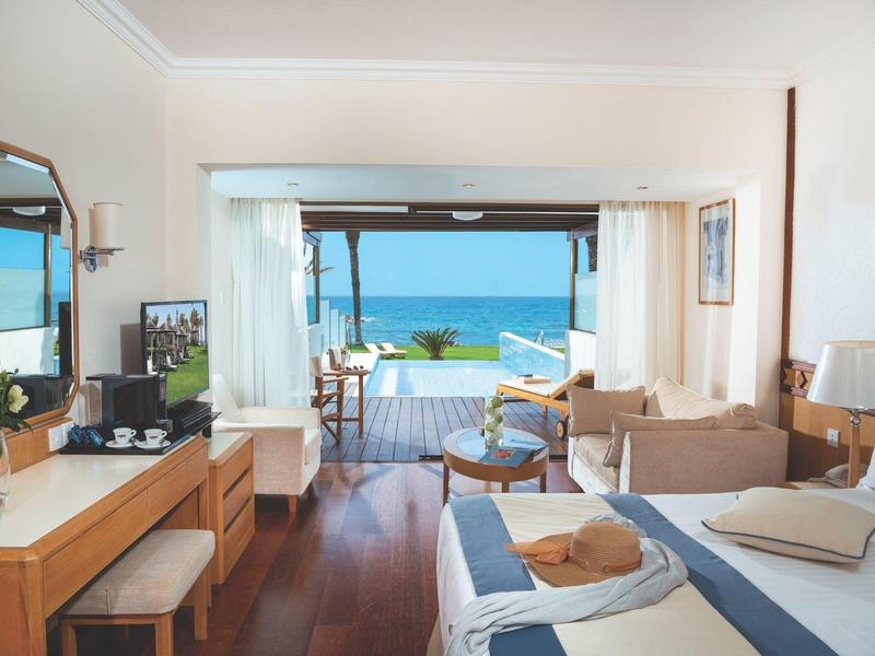 Helles Hotelzimmer mit Doppelbett, Sofa, Holzfußboden und Blick auf Meer durch große Fensterfront.