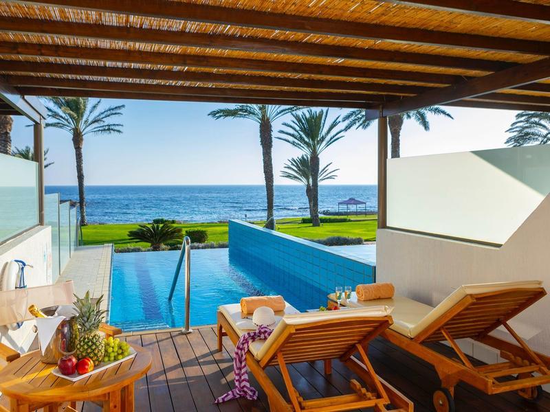 Blick von überdachter Terrasse mit Holzliegen auf blauen Pool, Palmen und Meer im Hintergrund bei sonnigem Himmel.