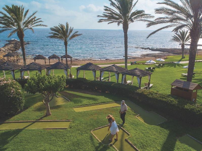 Grüner Garten mit Palmen, Pavillons und Minigolfplatz am Meer bei sonnigem Wetter.