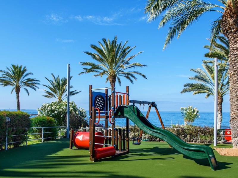 Spielplatz mit Rutsche, Klettergerüst und Palmen vor blauem Himmel und Meer.