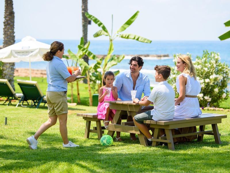 Familie sitzt an Holztisch im Garten, Kind bringt Essen, Meer und Sonnenschirm im Hintergrund.