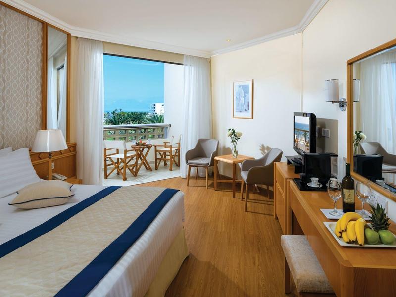 Helles Hotelzimmer mit Doppelbett, Balkon, zwei Stühlen, Schreibtisch und Fernseher, Holzboden.