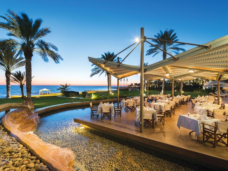 Terrasse mit gedeckten Tischen, Palmen und Wasserkanal bei Sonnenuntergang am Meer.