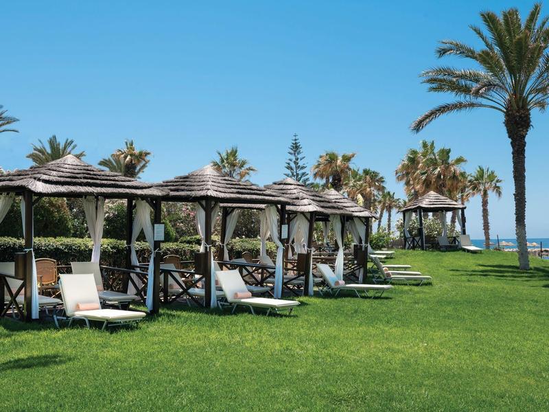 Grüne Wiese mit Liegestühlen unter Strohdächern und Palmen am blauen Himmel am Meer.