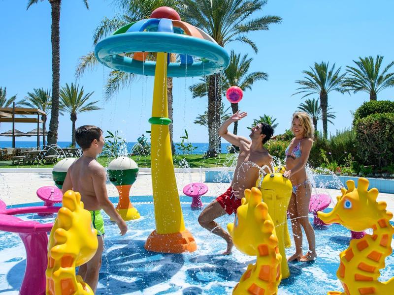 Kinder spielen fröhlich in einem bunten Wasserspielplatz mit gelben Seepferdchen und Palmen.