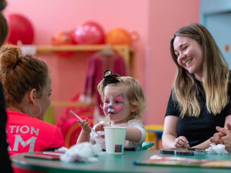 Kleines Mädchen mit Gesichtsbemalung sitzt am Tisch, umgeben von lächelnden Erwachsenen in hellem Raum.