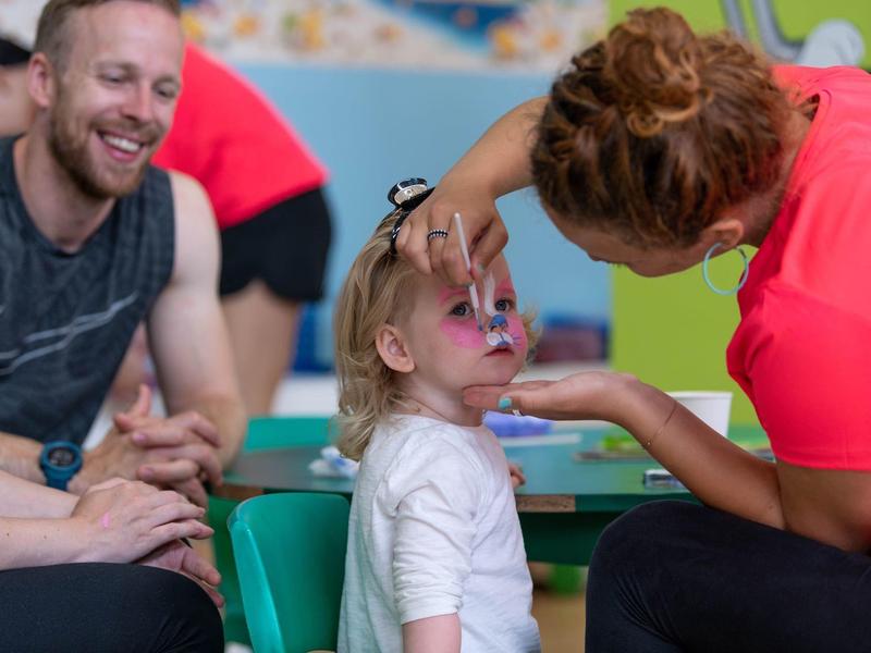 Ein Mädchen bekommt bunte Schmetterlingsgesichtsbemalung von einer Frau, Vater lächelt daneben.