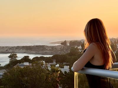 Frau mit roten Haaren blickt zum Sonnenuntergang über Küstenlandschaft und Häuser.